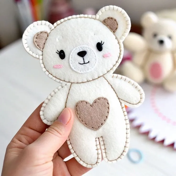 Finished white felt teddy bear with a beige heart on its tummy and rosy cheeks, held in a hand